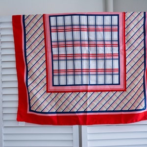 Può includere: Una sciarpa quadrata con bordo rosso e motivo a quadri bianco, rosa e blu. La sciarpa è appesa a una porta a persiane bianche. La sciarpa ha un motivo a quadri centrale e un motivo a quadri diagonale.