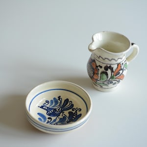 Può includere: Una piccola lattiera in ceramica e una ciotola abbinata. La lattiera presenta un motivo floreale dipinto a mano in arancione, verde e nero. La ciotola è decorata con un motivo a uccellino blu e bande circolari blu lungo il bordo interno ed esterno.