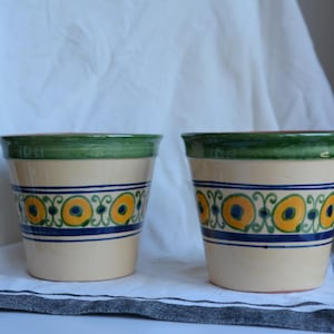 Op de afbeelding: Twee keramische bloempotten met een crèmekleurige basis. Elke pot heeft een groene rand en een blauw en geel decoratieve band. De potten zijn cilindrisch en staan op een wit en grijs gestreepte doek.