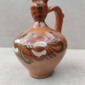 May include: A small, hand-painted terracotta jug with a rounded body and a handle. The jug features floral designs in white, brown, and green. The rim and handle are a darker shade of brown.