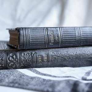 May include: Two antique, dark gray books stacked on a gray and white striped cloth. The top book has the word "Funeral" embossed on the spine. The bottom book has ornate floral and geometric designs.