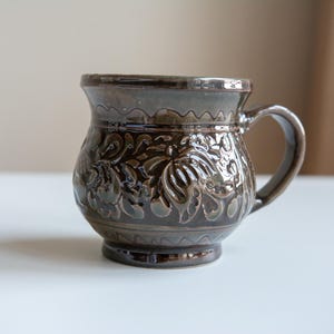 Può includere: Una tazza in ceramica marrone con manico, con un motivo floreale in rilievo dettagliato. La tazza ha un corpo arrotondato e un bordo leggermente svasato. La tazza è alta circa 10 cm.