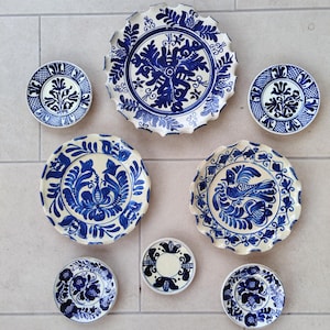 Puede incluir: Un conjunto de siete platos y platillos de cerámica con fondos de color crema y motivos intrincados de flores, hojas y aves pintados a mano en azul. El set incluye varios tamaños con bordes lisos y festoneados, mostrando patrones de arte popular tradicional en todas las piezas.