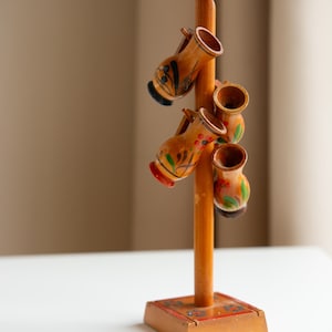May include: Wooden decorative stand with six small, hand-painted vases. The vases are orange with red, green, and black floral designs. The stand is brown with a square base and a central pole.