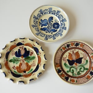 May include: Three round ceramic plates with cream-colored bases and hand-painted designs. One plate features blue floral patterns, another has a scalloped edge with blue, green, and brown floral motifs, and the third displays orange and blue flowers.