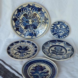 May include: A collection of hand-painted ceramic plates with a cream-colored base and intricate blue floral designs. The set includes plates of various sizes, showcasing traditional folk art patterns. The plates are arranged on a white cloth.