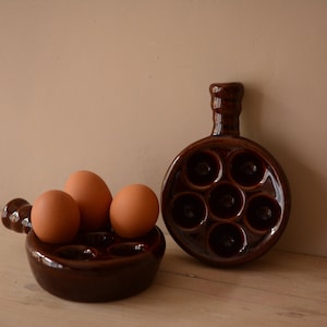 Puede incluir: Dos soportes para huevos de cerámica marrón. Un soporte tiene seis hendiduras individuales para huevos. El otro soporte tiene tres hendiduras y tres huevos están dentro.