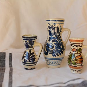 Può includere: Tre brocche in ceramica con manici, ognuna con un design unico. La brocca più grande presenta motivi floreali blu, mentre la più piccola ha un design floreale colorato. Le brocche sono su un panno bianco.