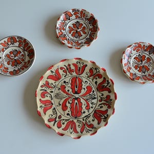 May include: A set of four ceramic plates featuring a cream-colored base with hand-painted red and black floral patterns. The collection includes one large scalloped-edge plate and three smaller plates, each decorated with symmetrical botanical motifs.