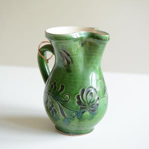 Op de afbeelding: Een groene keramieken kan met een glanzende afwerking, voorzien van een gebogen handvat en een uitlopende tuit. Het lichaam is versierd met donkergroene en zwarte handgeschilderde bloemmotieven en ranken. De binnenkant is wit.