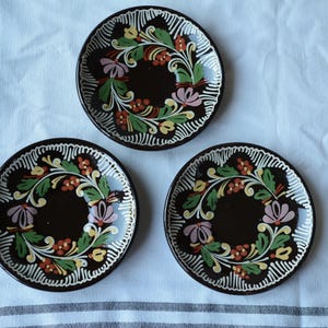 May include: Three round ceramic plates with a dark brown base color. Each plate features a hand-painted floral wreath design in shades of green, yellow, red, and light purple, accented by white linear patterns along the outer rim of the plates.