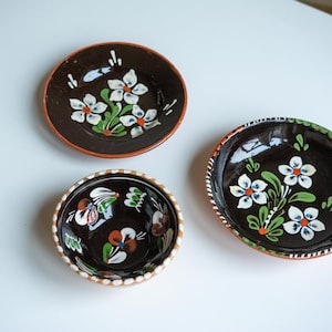 May include: Three hand-painted ceramic plates with floral designs. The plates are dark brown with white, green, and orange floral patterns. The rims are decorated with orange and white accents. A small bowl is also included.