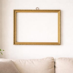Könnte beinhalten: Ein rechteckiger, verzierter goldener Bilderrahmen hängt an einer weißen Wand. Der Rahmen hat detaillierte Schnitzereien und einen dekorativen Rand. Im Vordergrund sind ein Sofa und eine Pflanze zu sehen. Der Rahmen ist leer.