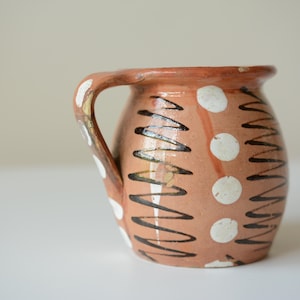May include: A terracotta-colored ceramic mug with a single handle, featuring a hand-painted pattern of vertical dark brown zigzag lines alternating with columns of solid white dots that wrap around the rounded body of the vessel.