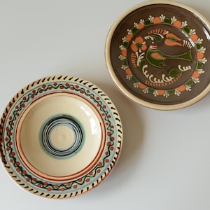 May include: Two round ceramic plates. One plate is cream-colored with blue and red concentric circles and a decorative border. The other plate is brown with a central green bird illustration surrounded by orange and green floral patterns.