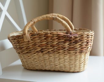 May include: A woven basket with two arched handles. The basket is made of natural fibers in shades of tan and brown. A small, decorative bow is attached near the handles. The basket is sitting on a white surface.