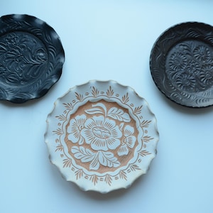 May include: Three round decorative plates with scalloped edges and embossed floral patterns. Two plates are solid black, while the central plate features a white rim and a textured, light brown center with a prominent white floral design.