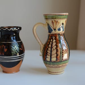 Puede incluir: Dos jarras de cerámica con asa. La jarra de la izquierda es negra con líneas horizontales blancas y patrones en zigzag naranjas y verdes. La jarra de la derecha es de color crema con patrones verticales en forma de hoja marrones y verdes.