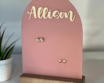 May include: A pink acrylic earring holder with a wooden base. The holder has a rounded arch shape with the name "Allison" written in gold lettering at the top. There are several holes for earrings on the front of the holder.
