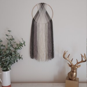 Puede incluir: Un adorno de pared con un anillo de metal dorado y hebras verticales de hilo en tonos grises, blanco y negro. La obra de arte est&aacute; montada en una pared blanca. Una estatua de cabeza de ciervo dorada y una jarra blanca con vegetaci&oacute;n est&aacute;n sobre una superficie de madera.