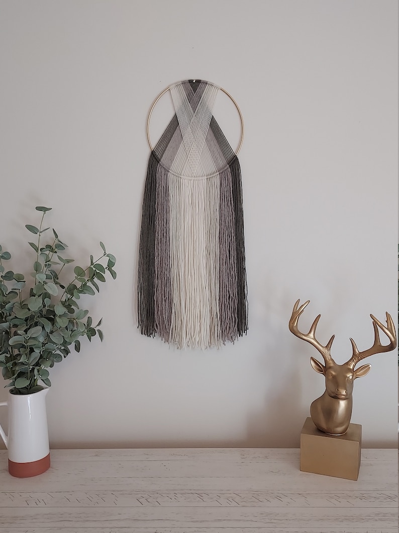 Puede incluir: Un adorno de pared con un anillo de metal dorado y hebras verticales de hilo en tonos grises, blanco y negro. La obra de arte est&aacute; montada en una pared blanca. Una estatua de cabeza de ciervo dorada y una jarra blanca con vegetaci&oacute;n est&aacute;n sobre una superficie de madera.