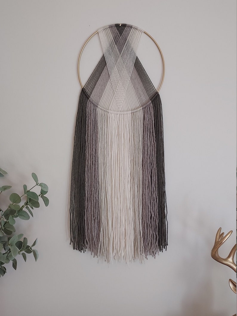 Puede incluir: Un tapiz de pared hecho a mano con un anillo de metal dorado y un dise&ntilde;o tejido en tonos grises, blancos y negros. Los hilos de lana caen en cascada, creando una est&eacute;tica texturizada y moderna. Una pieza elegante para la decoraci&oacute;n del hogar.