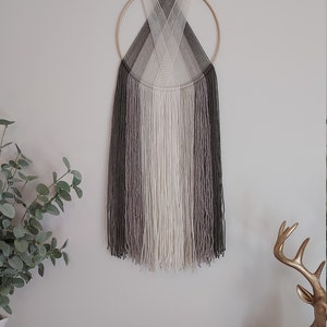 Puede incluir: Un adorno de pared con un anillo de metal dorado y hebras verticales de hilo en tonos grises, blancos y negros. El hilo est&aacute; dispuesto en un patr&oacute;n de gradiente, creando un dise&ntilde;o moderno y minimalista. Esta pieza decorativa a&ntilde;ade textura e inter&eacute;s visual a cualquier espacio.