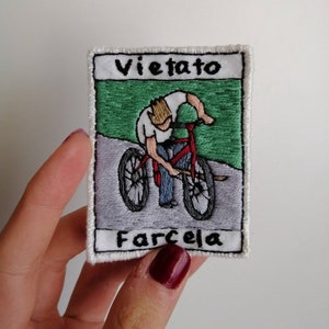 Può includere: Toppa ricamata raffigurante un ciclista su una bicicletta rossa. La toppa ha un bordo bianco con le parole "Vietato" e "Farcela" in nero. Lo sfondo mostra un'area verde e una strada grigia.