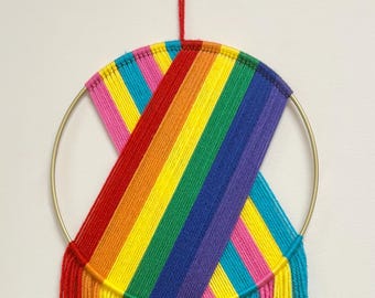 May include: A vibrant rainbow-colored wall hanging. The artwork features a gold metal ring with a woven design in red, orange, yellow, green, blue, purple, and pink. Below the ring, the colors cascade downward in a fringe of yarn.