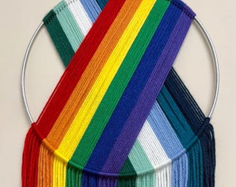 May include: A colorful rainbow wall hanging. The artwork features a metal ring with a woven design in a spectrum of colors, including red, orange, yellow, green, blue, and purple. The bottom of the piece has long, fringed yarn strands.