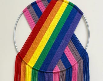 May include: A rainbow-colored yarn wall hanging. The design features a silver metal hoop with yarn woven across in a rainbow pattern. Vertical yarn strands hang below, creating a textured, colorful display. The colors include red, orange, yellow, green, blue, purple, pink, and gray.