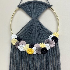 May include: A gray yarn dream catcher with a gold metal hoop and a fringe of yarn. The hoop is decorated with a row of felt flowers in shades of yellow, white, black, and gray.
