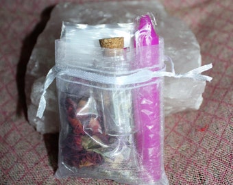 Può includere: Una borsa di organza bianca piena di petali di rosa secchi, una piccola bottiglia di vetro con un tappo di sughero e una candela viola. La borsa è legata con un nastro bianco.