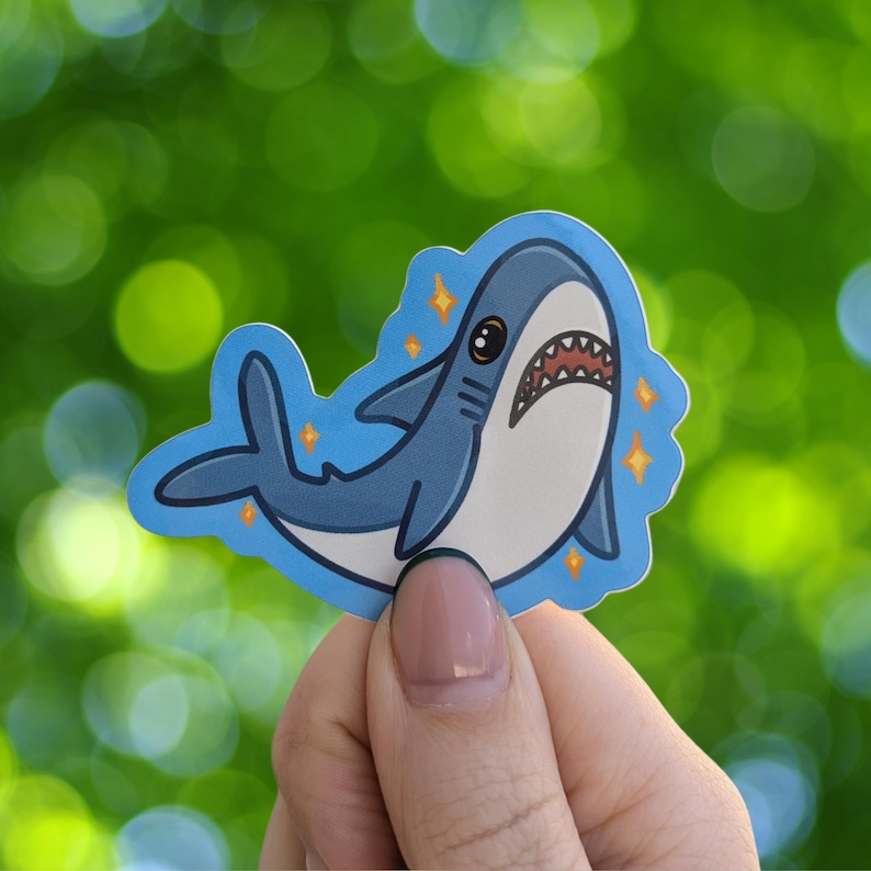 Puede incluir: Una pegatina de tibur&oacute;n de dibujos animados azul y blanco con detalles brillantes. El tibur&oacute;n tiene la boca muy abierta y ojos grandes.