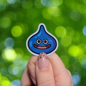 Op de afbeelding: Een blauwe, druppelvormige sticker met een lachend gezicht en witte omtrek. De sticker heeft twee zwarte ogen en een roze mond. De achtergrond is een wazig groen met bokeh-effect.