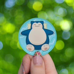 Op de afbeelding: Een ronde sticker met een cartoon Snorlax-personage in blauw en crème, rustend op een lichtblauwe ondergrond met golfpatronen. De sticker heeft een witte rand en staat tegen een vage groene achtergrond.