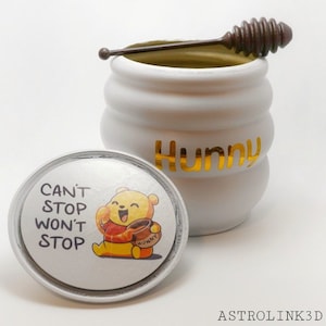 Może przedstawiać: Biały ceramiczny słoik na miód z postacią Kubusia Puchatka na pokrywce. Pokrywka ma napis "Can't Stop Won't Stop". Słoik ma napis "Hunny" złotymi literami. W zestawie znajduje się brązowa łyżeczka do miodu.