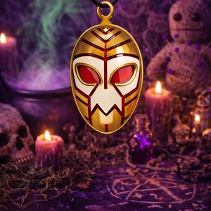 May include: Gold-colored pendant with a mask design, featuring red and white accents, suspended from a black cord. The pendant is oval-shaped with a gold bail. The background includes candles, skulls, and a voodoo doll, creating a mystical theme.