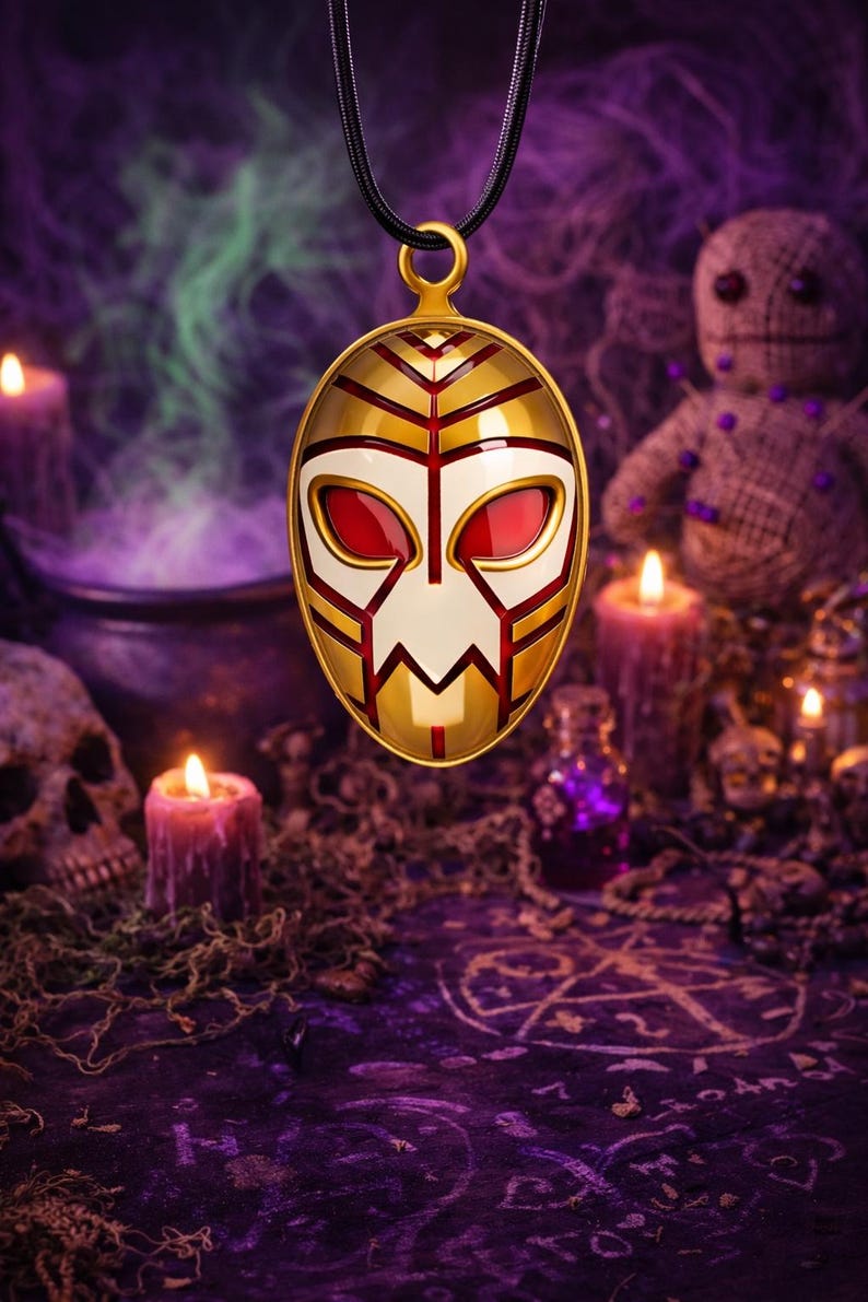 May include: Gold-colored pendant with a mask design, featuring red and white accents, suspended from a black cord. The pendant is oval-shaped with a gold bail. The background includes candles, skulls, and a voodoo doll, creating a mystical theme.