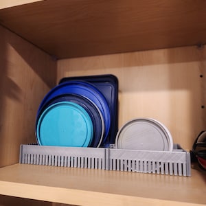 May include: A gray plastic shelf organizer with blue and turquoise plastic lids stacked on top. The organizer has a series of vertical slots to hold the lids.