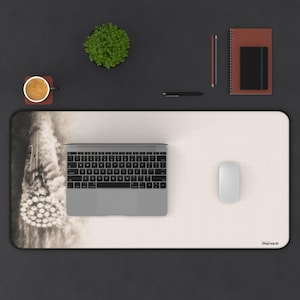Puede incluir: Una alfombrilla de escritorio rectangular con un patrón de cuadrícula y una ilustración en blanco y negro de un lanzamiento de cohete a la izquierda. La alfombrilla incluye un portátil plateado, un ratón blanco, una taza, una planta y un cuaderno.