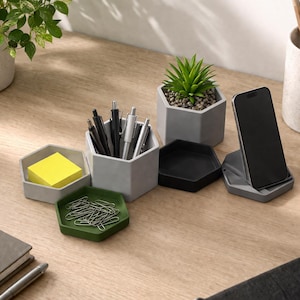 Pode incluir: Uma coleção de organizadores de mesa hexagonais em várias cores, incluindo cinza, preto e verde. Os organizadores contêm itens como canetas, notas adesivas, clipes de papel e um smartphone. Pequenas plantas em vasos e um porta-canetas também estão presentes na mesa de madeira.