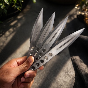 以下が含まれることがあります： 鋭い刃と柄に丸い穴がある、銀色の手裏剣のような投げナイフが5本。手で持ち、グレーの背景に影が落ちている。