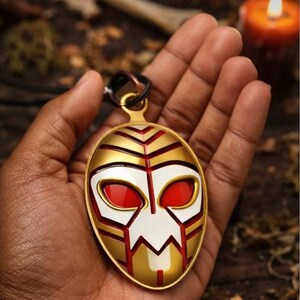 May include: Gold-colored pendant with a mask design. The mask features red eyes, white and gold accents, and a black cord. The pendant is held in a hand, with a blurred background including a candle and foliage.