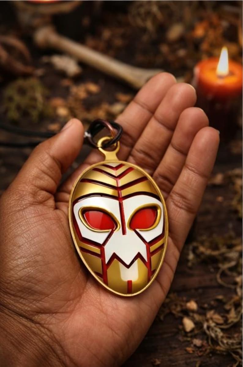 May include: Gold-colored pendant with a mask design. The mask features red eyes, white and gold accents, and a black cord. The pendant is held in a hand, with a blurred background including a candle and foliage.