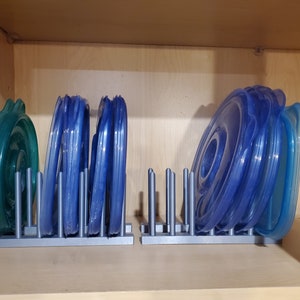 Puede incluir: Dos organizadores de tapas de plástico gris con tapas de plástico azul apiladas en su interior. Los organizadores están en un estante de madera.