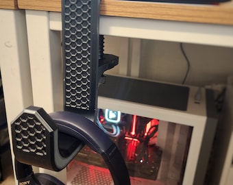 May include: Black headphone stand with a honeycomb pattern, holding a pair of black Sony headphones. The stand is clamped to a desk, with a computer visible in the background. The headphones have gold lettering.