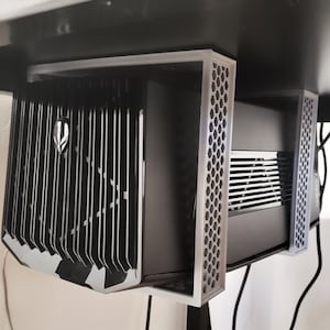 Könnte beinhalten: Ein schwarzes Computergehäuse mit einem wabenförmigen Metallrahmen. Das Gehäuse hat einen großen, schwarzen Lüfter an der Seite und einen kleineren Lüfter an der Vorderseite. Das Gehäuse ist mit einer Metallhalterung an einem Schreibtisch befestigt.