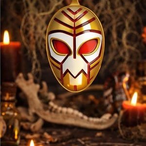 May include: Gold-colored pendant with a mask design, featuring red, white, and gold accents. The pendant hangs from a black cord. The mask has a stylized face shape. The background includes candles and a dark, textured surface.