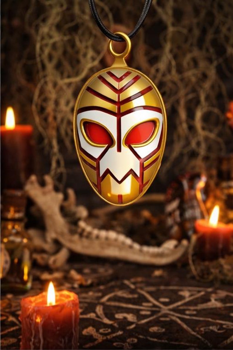 May include: Gold-colored pendant with a mask design, featuring red, white, and gold accents. The pendant hangs from a black cord. The mask has a stylized face shape. The background includes candles and a dark, textured surface.